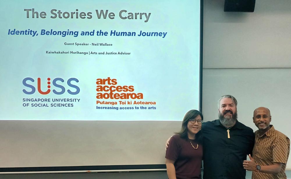 Jiunwen Wang, Neil Wallace and Siva Gopal Thaiyalan stand in front of a large screen before Neil's keynote. It has Arts Access Aotearoa's and SUSS's logos and has the title of Neil's keynote